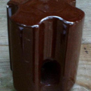 May include: A brown ceramic insulator with a glossy finish. The cylindrical shape has a distinctive four-lobed top and a central hole. The insulator is designed for electrical applications and is likely vintage.