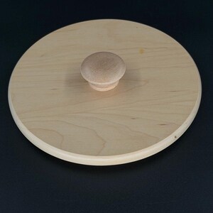 Puede incluir: Una tapa redonda de madera clara con un pequeño pomo redondeado en el centro. La tapa tiene una superficie lisa y un borde ligeramente más oscuro y redondeado. El pomo también es de madera clara.