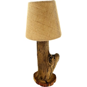 May include: A rustic table lamp featuring a burlap shade atop a natural wood base. The lamp's base is crafted from a weathered tree trunk, showcasing the wood's natural grain and texture. The shade is a light tan color.