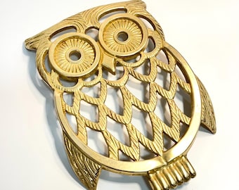 May include: A gold-colored, owl-shaped trivet. The trivet features a detailed design with openwork feathers and large, circular eyes. The owl's feet are visible at the bottom. The trivet is likely made of metal and is designed to protect surfaces from hot items.