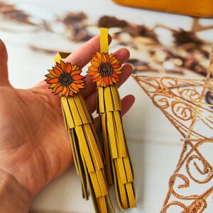 May include: Two yellow leather tassels with sunflower charms. The tassels are made of multiple layers of leather strips and have a frayed edge. The sunflowers are orange and yellow with brown centers.