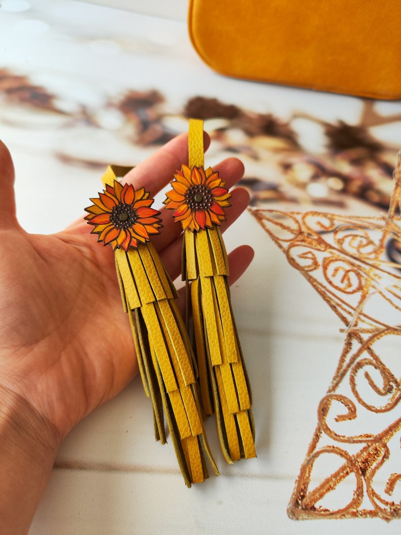 May include: Two yellow leather tassels with sunflower charms. The tassels are made of multiple layers of leather strips and have a frayed edge. The sunflowers are orange and yellow with brown centers.