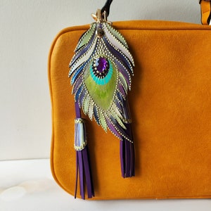 May include: A mustard yellow leather handbag with a purple and green peacock feather charm. The charm has a purple gemstone in the center and gold accents.