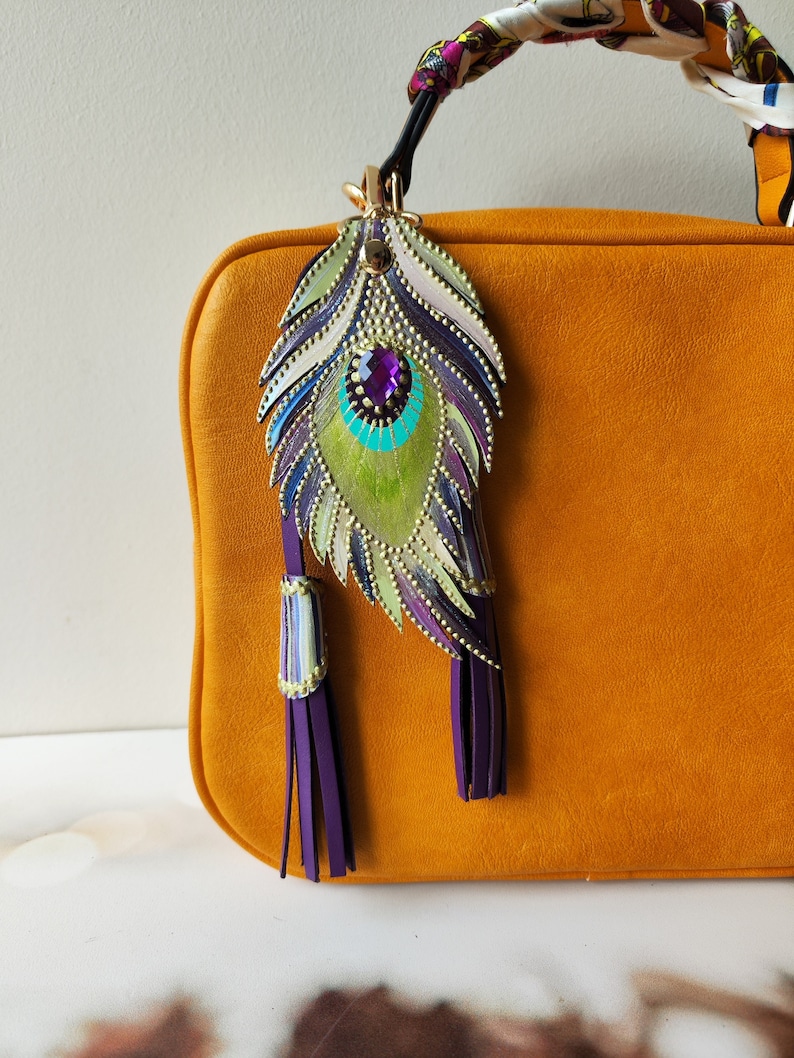 May include: A mustard yellow leather handbag with a purple and green peacock feather charm. The charm has a purple gemstone in the center and gold accents.