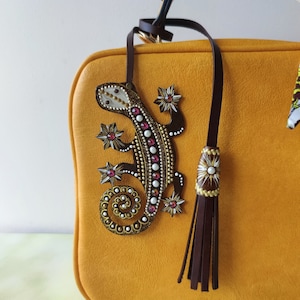 Könnte beinhalten: Eine senfgelbe Ledertasche mit einem braunen Lederriemen und einem Quastenanhänger. Die Handtasche hat einen dekorativen Eidechsen-Anhänger mit goldenen und weißen Akzenten und rosafarbenen Strasssteinen.