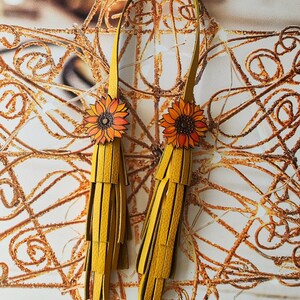 May include: A pair of yellow leather tassel earrings with sunflower accents. The earrings are hanging from a gold background.
