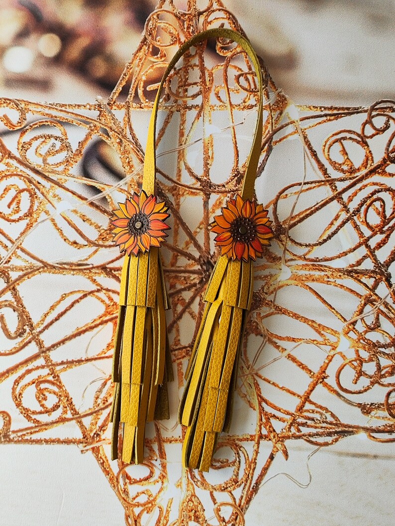 May include: A pair of yellow leather tassel earrings with sunflower accents. The earrings are hanging from a gold background.