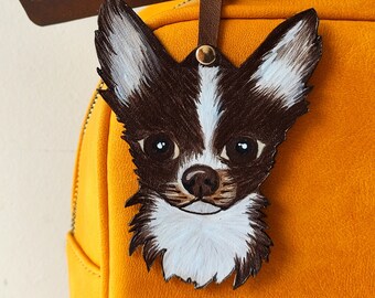 May include: A brown leather handbag with a painted chihuahua face charm. The charm is attached to the bag with a brown leather strap.