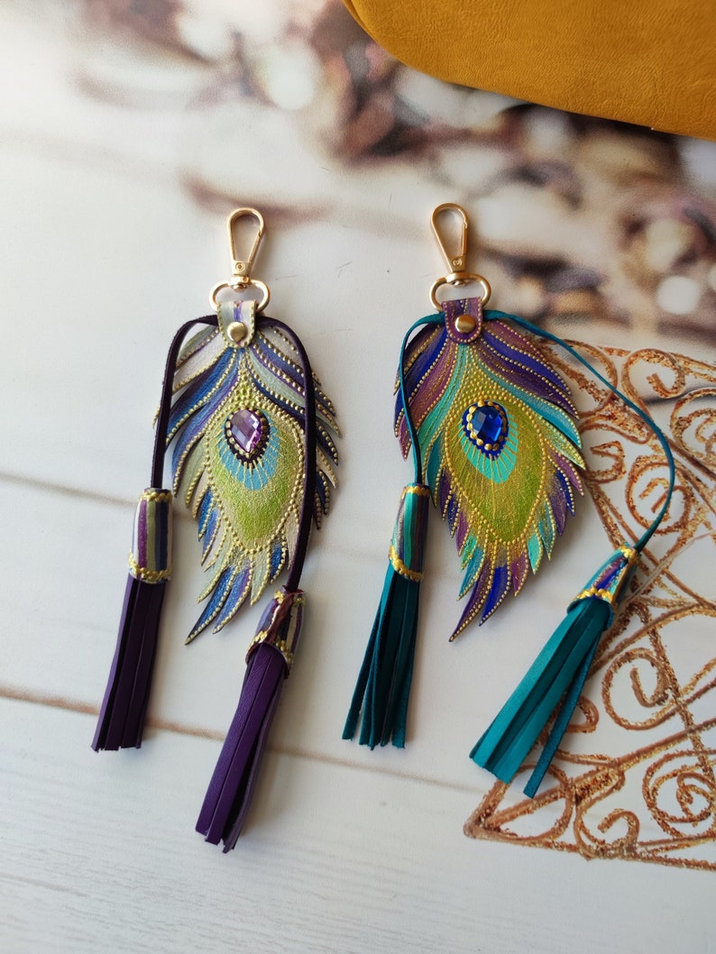 May include: Two peacock feather keychains with gold accents and jeweled eyes. One keychain has purple tassels and the other has teal tassels.