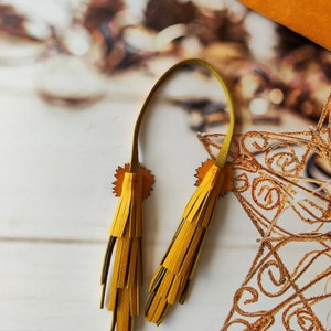 May include: Two yellow leather tassels with a green leather cord. The tassels are attached to a piece of leather with a decorative design.