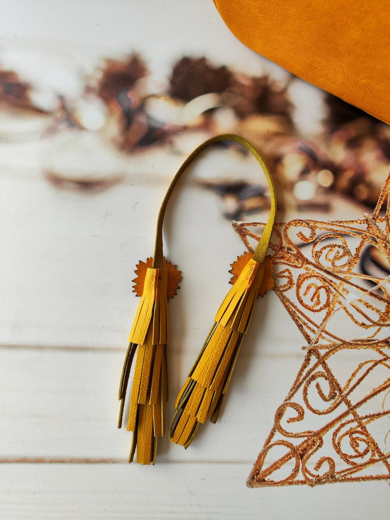 May include: Two yellow leather tassels with a green leather cord. The tassels are attached to a piece of leather with a decorative design.