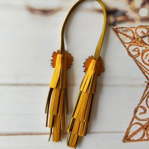 May include: A pair of yellow leather tassels with a green leather strap. The tassels are attached to the strap with a small, circular, decorative piece.