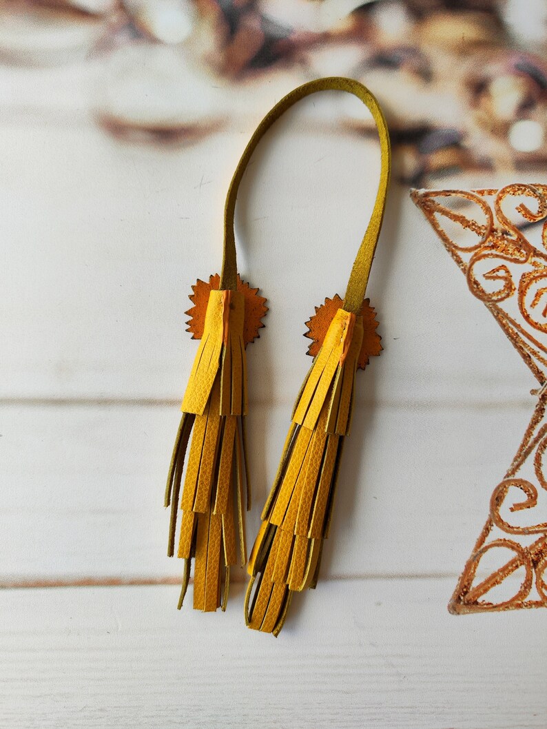 May include: A pair of yellow leather tassels with a green leather strap. The tassels are attached to the strap with a small, circular, decorative piece.