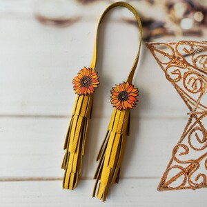 May include: A pair of yellow leather tassels with sunflower embellishments. The tassels are attached to a yellow leather strap.