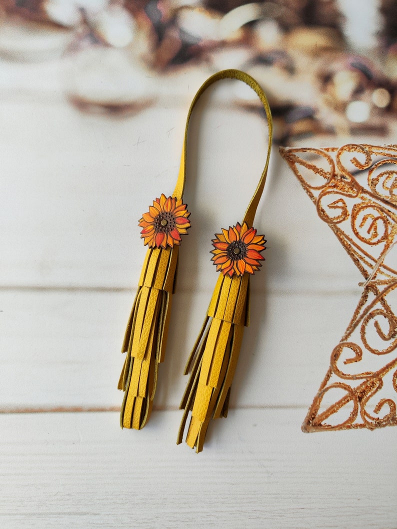 May include: A pair of yellow leather tassels with sunflower embellishments. The tassels are attached to a yellow leather strap.