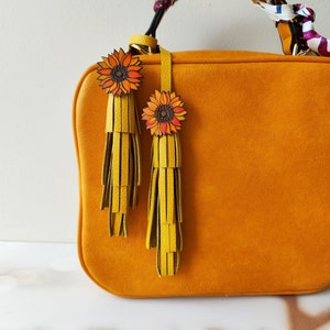 May include: A mustard yellow handbag with a brown leather strap and two yellow tassels with sunflower charms.