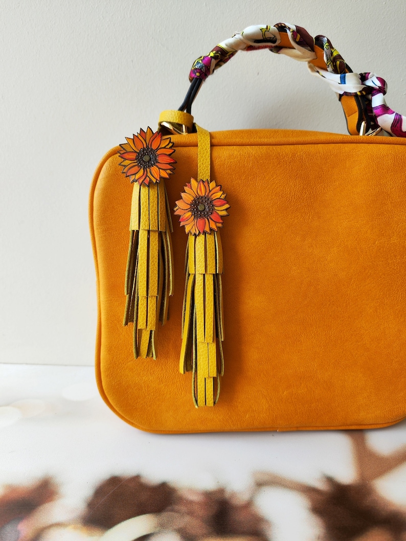 May include: A mustard yellow handbag with a brown leather strap and two yellow tassels with sunflower charms.
