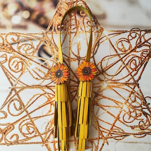 May include: A pair of yellow leather tassel earrings with sunflower accents. The earrings are hanging from a gold star-shaped background.