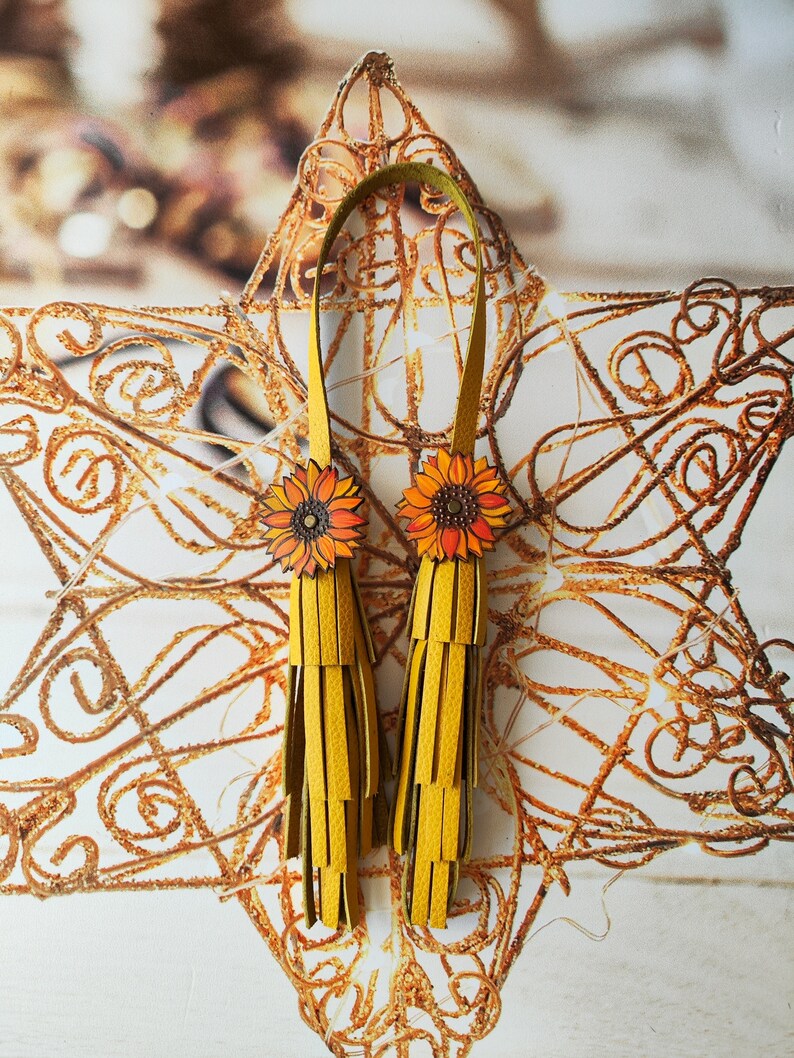 May include: A pair of yellow leather tassel earrings with sunflower accents. The earrings are hanging from a gold star-shaped background.