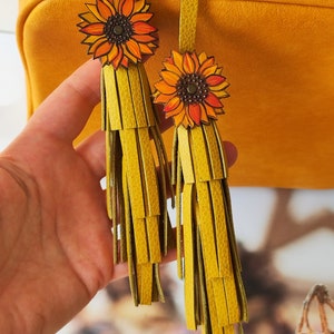 May include: Two yellow leather tassels with sunflower charms. The tassels are made of multiple layers of leather strips and the sunflowers are painted with orange and yellow petals and a brown center.
