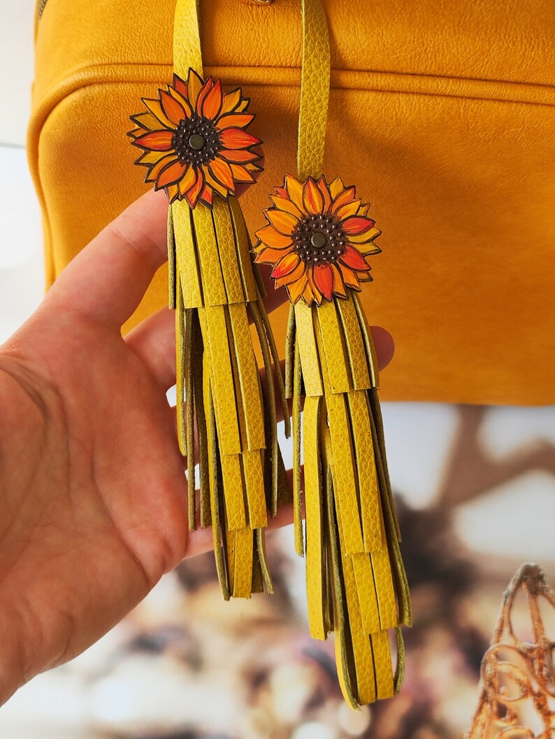May include: Two yellow leather tassels with sunflower charms. The tassels are made of multiple layers of leather strips and the sunflowers are painted with orange and yellow petals and a brown center.