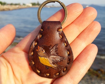 May include: A brown leather keychain with a stitched edge and a painted image of a yellow bird with brown wings.