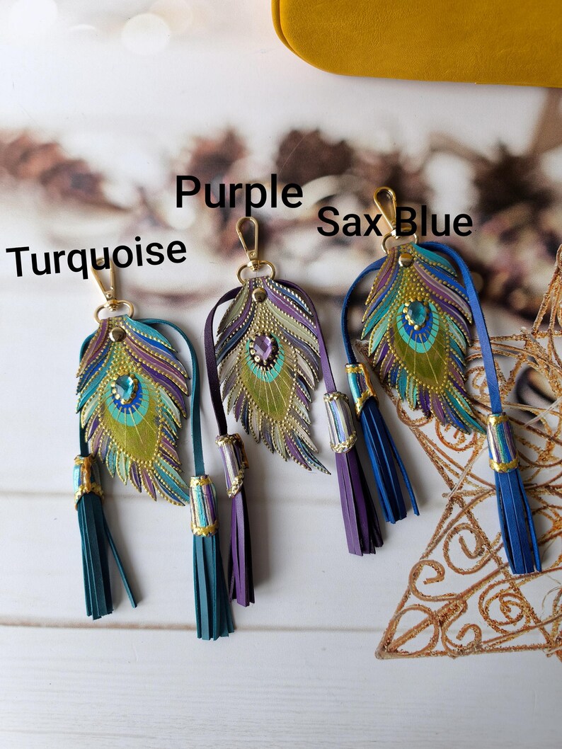 May include: Four peacock feather keychains with tassels. The feathers are made of leather and have a gold, green, blue, and purple color scheme. The tassels are in turquoise, purple, blue, and sax blue.