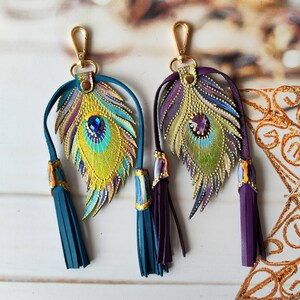 May include: Two gold-tone keychains with peacock feather charms. The feathers are painted with gold, green, blue, and purple accents. Each keychain has a tassel, one is teal and the other is purple.