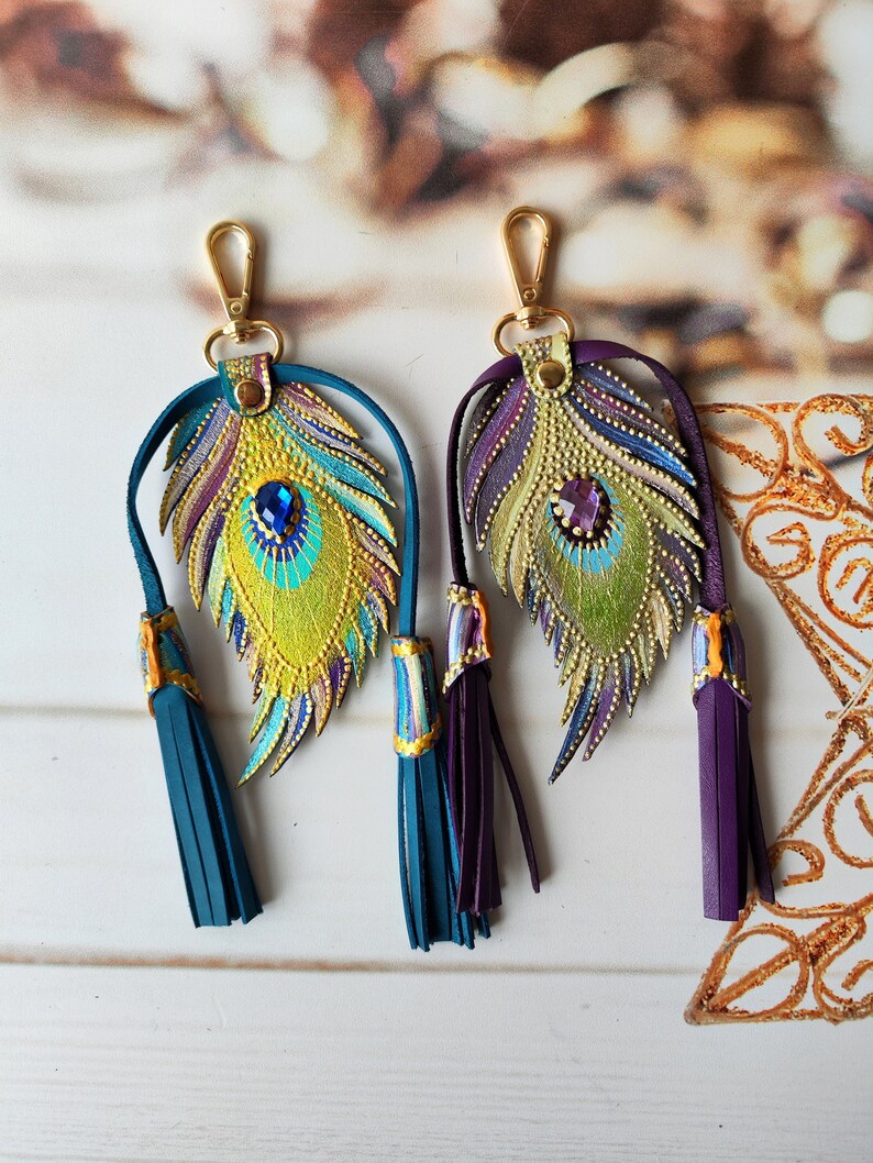May include: Two gold-tone keychains with peacock feather charms. The feathers are painted with gold, green, blue, and purple accents. Each keychain has a tassel, one is teal and the other is purple.
