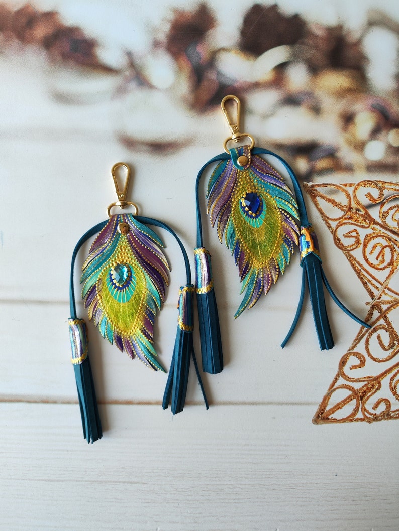 May include: Two peacock feather-shaped keychains with gold accents and blue tassels. The feathers are painted with green, yellow, purple, and blue colors and have a blue rhinestone in the center.