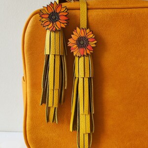 May include: A mustard yellow leather handbag with two sunflower-shaped leather tassels. The tassels are yellow and green and have a fringe detail.