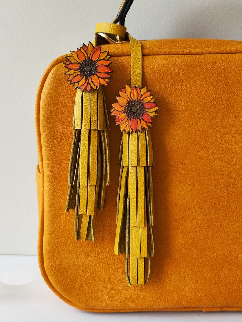 May include: A mustard yellow leather handbag with two sunflower-shaped leather tassels. The tassels are yellow and green and have a fringe detail.