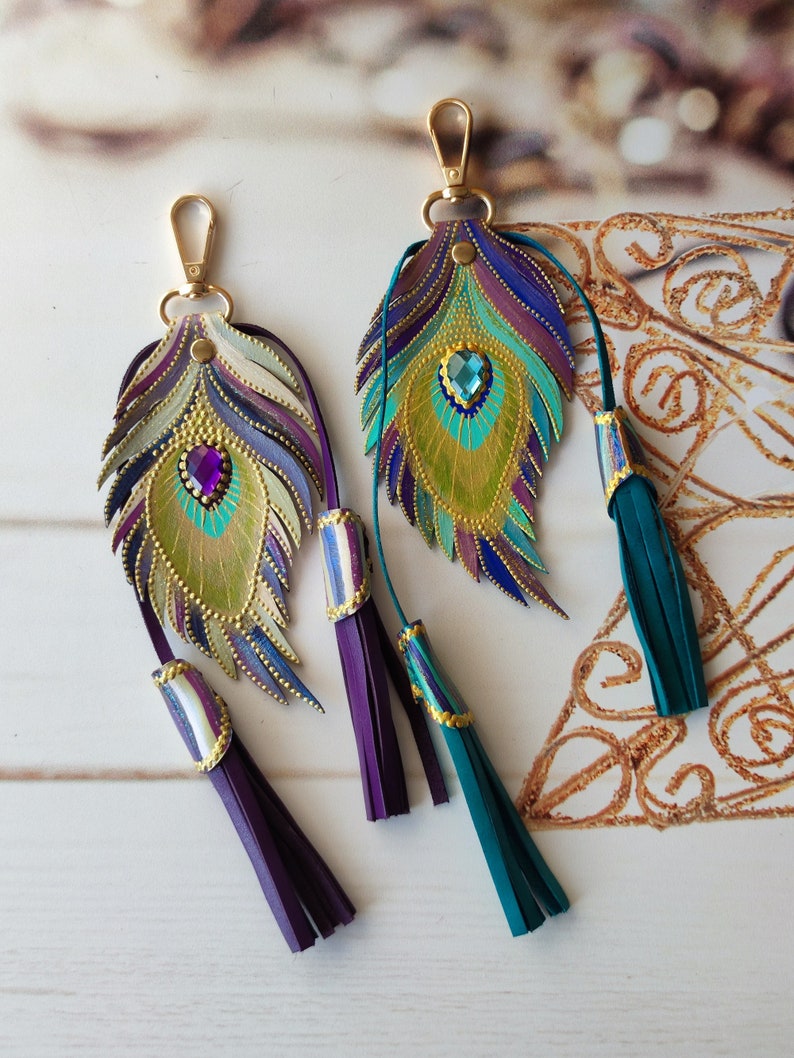 May include: Two leather peacock feather keychains with gold accents and jeweled eyes. Each keychain has a purple or teal tassel.