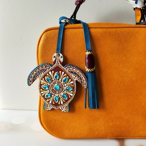 Puede incluir: Un bolso de mano de cuero naranja brillante con un borla azul y marrón. Un pequeño amuleto de tortuga pintado a mano con detalles en turquesa y oro cuelga del bolso.