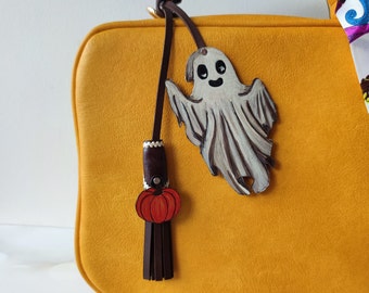 Puede incluir: Un bolso de mano de cuero amarillo con un amuleto de fantasma y calabaza. El amuleto de fantasma es blanco con ojos negros y una sonrisa. El amuleto de calabaza es naranja con un tallo verde.
