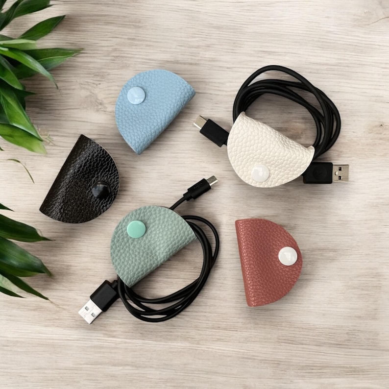 May include: Five leather cord organizers in different colors: light blue, black, light green, beige, and brown. Each organizer has a button closure and is holding a black cord.