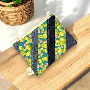 May include: A small, quilted pouch with a cactus print and denim accents. The pouch features a teal background with yellow cacti and pink flowers. Denim stripes and a gold zip complete the design. The label reads "Itsy Bitsy Stitches."