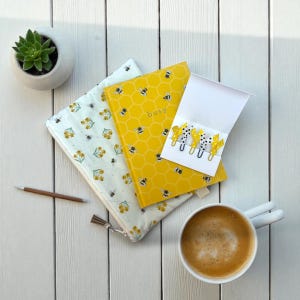 May include: A flat lay composition with a yellow notebook featuring a honeycomb design and the word "busy", a floral pouch, and a white card with bee-shaped paper clips. A coffee cup and a small succulent in a white pot are also visible.