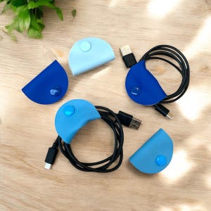 May include: Four blue and light blue leather cable organisers with a button closure. The organisers are on a wooden surface.