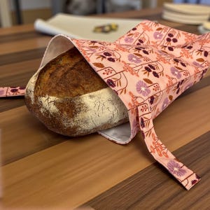 May include: A round loaf of bread rests inside a pink floral bread bag. The bag features a repeating pattern of purple and burgundy flowers and stems. The bread has a golden-brown crust and is partially visible.