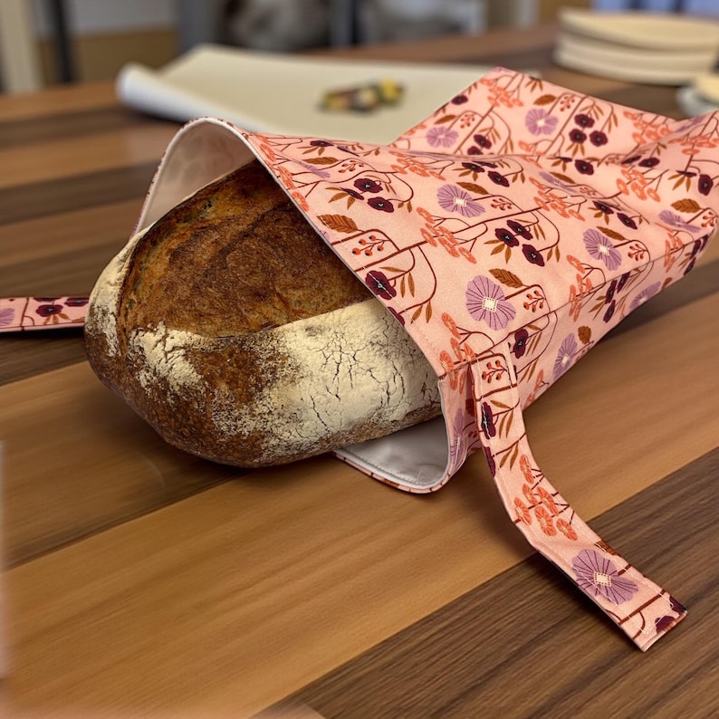May include: A round loaf of bread rests inside a pink floral bread bag. The bag features a repeating pattern of purple and burgundy flowers and stems. The bread has a golden-brown crust and is partially visible.