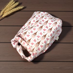 May include: A white fabric lunch bag featuring a repeating pattern of pink flowers and brown stems. The bag has a roll-top closure with a handle and a small rectangular tag on the side that reads The Little Stitcher.