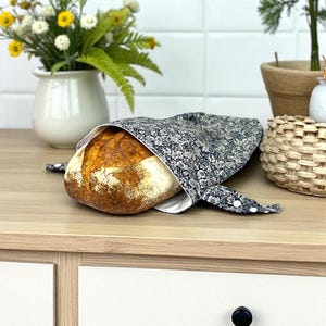 May include: A round loaf of bread partially enclosed in a navy blue floral bread bag. The bag is open, revealing the crusty, golden-brown bread. The bag has white lining and is tied with white buttons.
