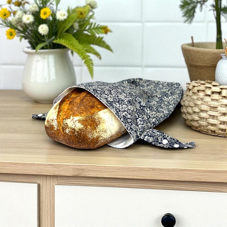 May include: A round loaf of bread partially enclosed in a navy blue floral bread bag. The bag is open, revealing the crusty, golden-brown bread. The bag has white lining and is tied with white buttons.