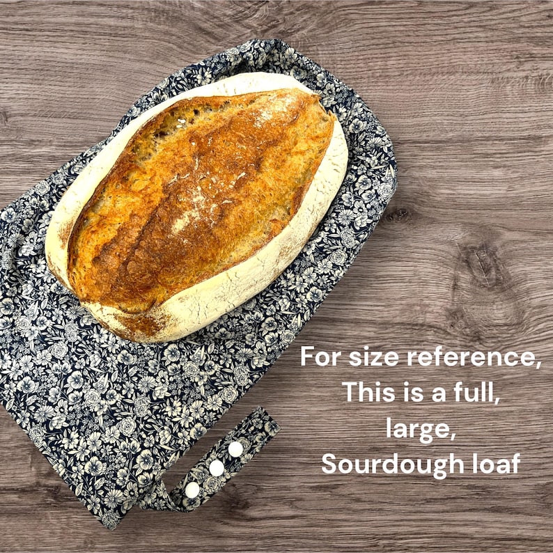 May include: A large, golden-brown sourdough loaf rests on a navy blue floral patterned bread basket. The basket has a handle with three white buttons. Text on the image reads: "For size reference, This is a full, large, Sourdough loaf."