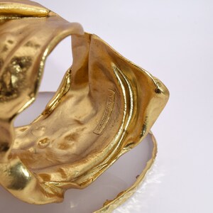 May include: A gold-colored cuff bracelet with a textured, organic design. The bracelet is resting on a white and gold-edged surface. The text "KARINE SULTAN" is visible on the bracelet.