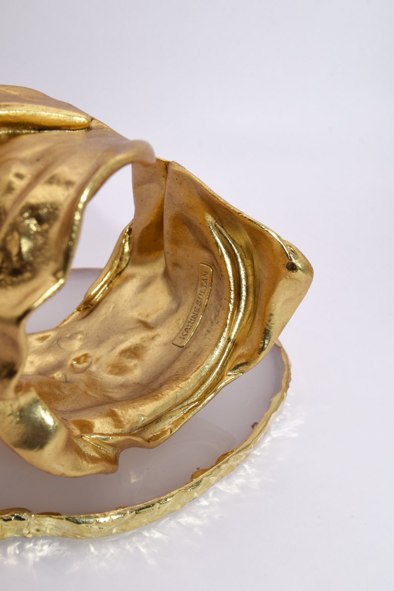 May include: A gold-colored cuff bracelet with a textured, organic design. The bracelet is resting on a white and gold-edged surface. The text "KARINE SULTAN" is visible on the bracelet.