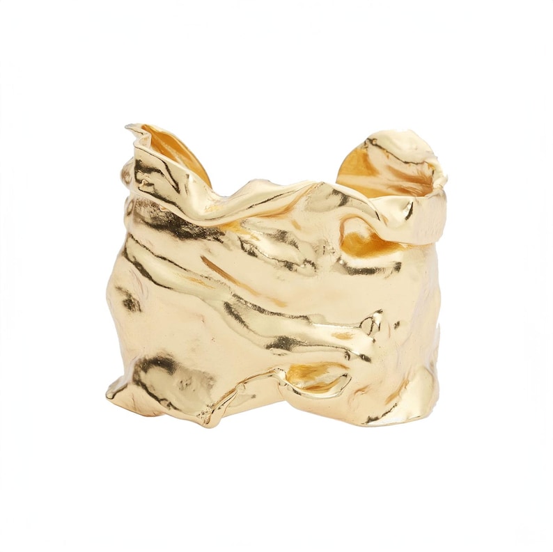 May include: A gold-colored cuff bracelet with a textured, crumpled appearance. The metal appears to be molded with folds and ridges, creating a unique, abstract design. The bracelet is set against a white background.