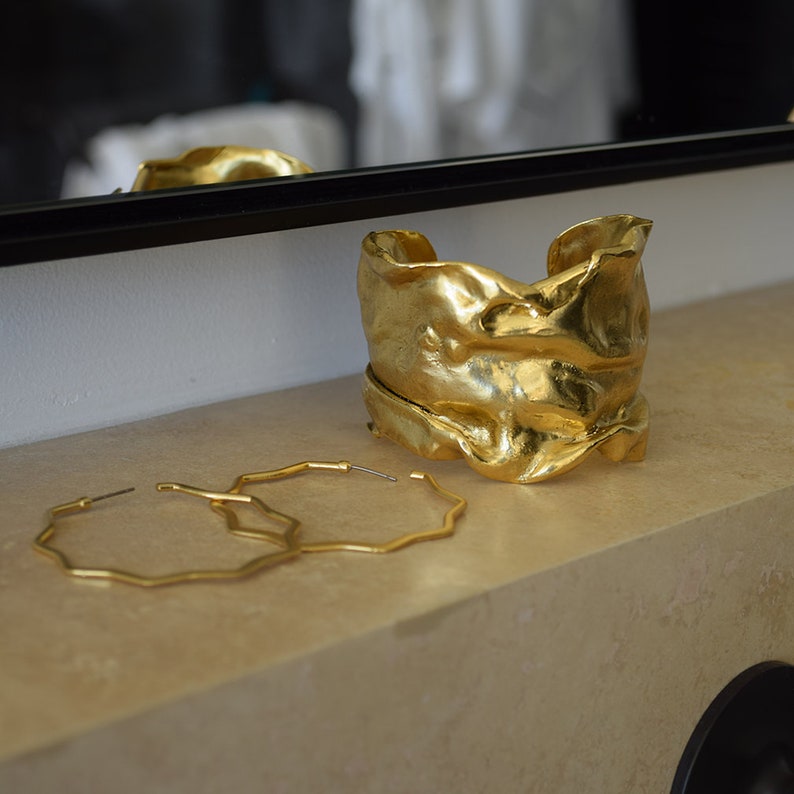 May include: A gold-colored cuff bracelet with a textured, crumpled appearance sits next to a pair of gold hoop earrings with a wavy design. The jewelry is displayed on a light-colored surface, with a mirror in the background.