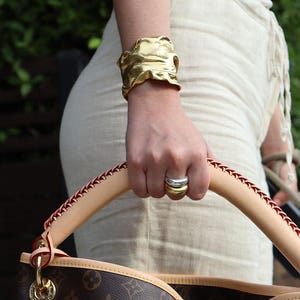 May include: A gold-colored cuff bracelet with a textured, organic design. The bracelet is worn on a wrist, paired with two rings, one gold and one silver. The person is holding a handbag with a brown and tan pattern.